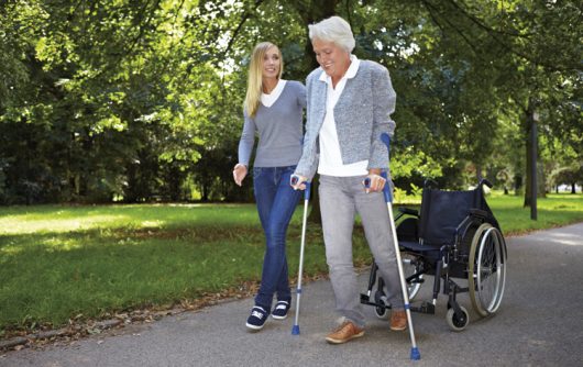 senior woman and young woman walking in park. Senior woman uses wheelchair and arm braces