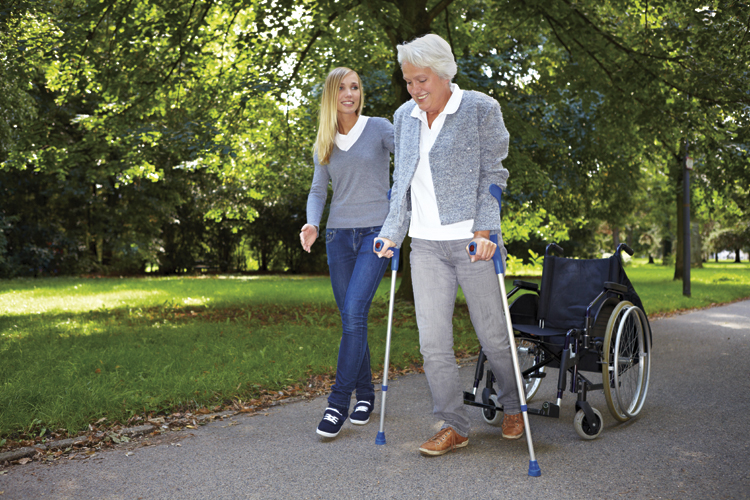 senior woman and young woman walking in park. Senior woman uses wheelchair and arm braces