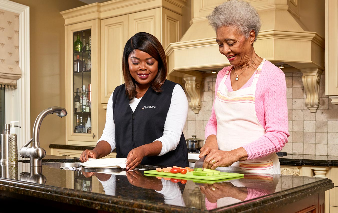 Caregiver providing assistance in the kitchen to an old woman who is cutting vegetables with a knife.