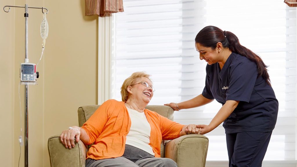 Nurse holding senior woman's hand. The senior woman is sat on the sofa while the nurse is standing. They both are smiling at each other.