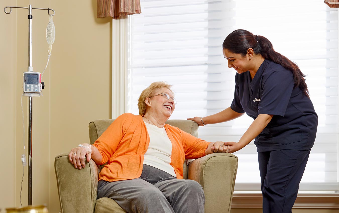 Nurse holding senior woman's hand. The senior woman is sat on the sofa while the nurse is standing. They both are smiling at each other.