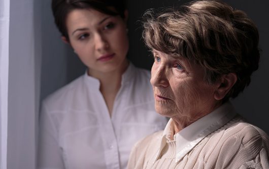 senior woman looking sad out window with young woman looking at her with concern