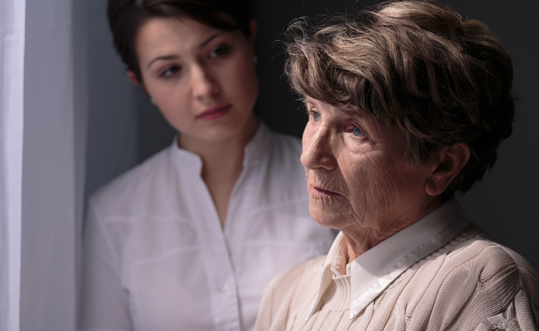 senior woman looking sad out window with young woman looking at her with concern