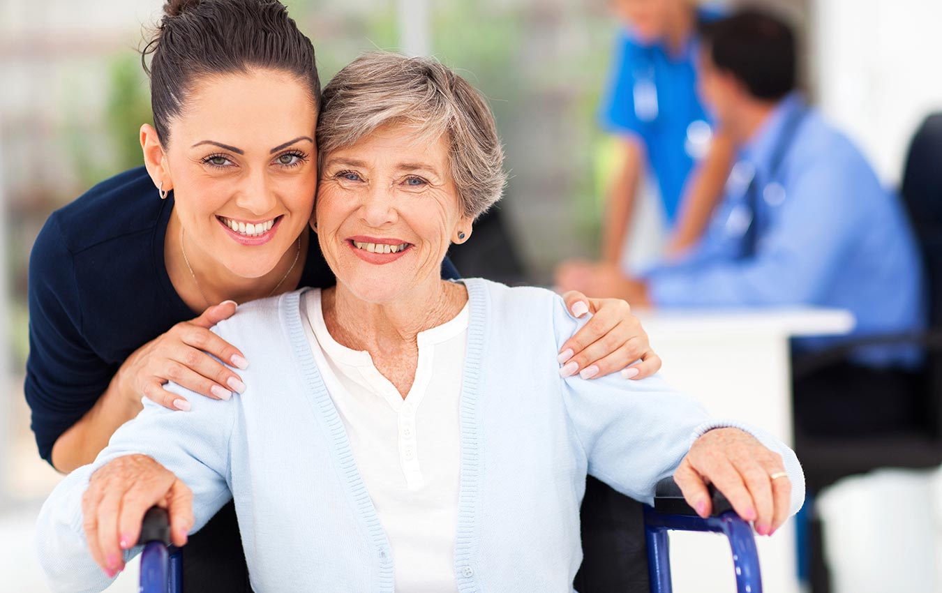 Nurse comforting a senior woman sitting on a chair. They both are smiling for a photograph.