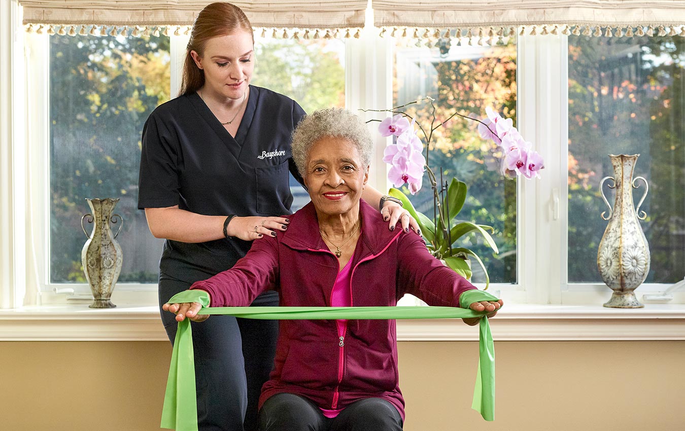 A caregiver assisting a senior woman with physiotherapy. The woman is sat holding a resistance band trying to perform an exercise.