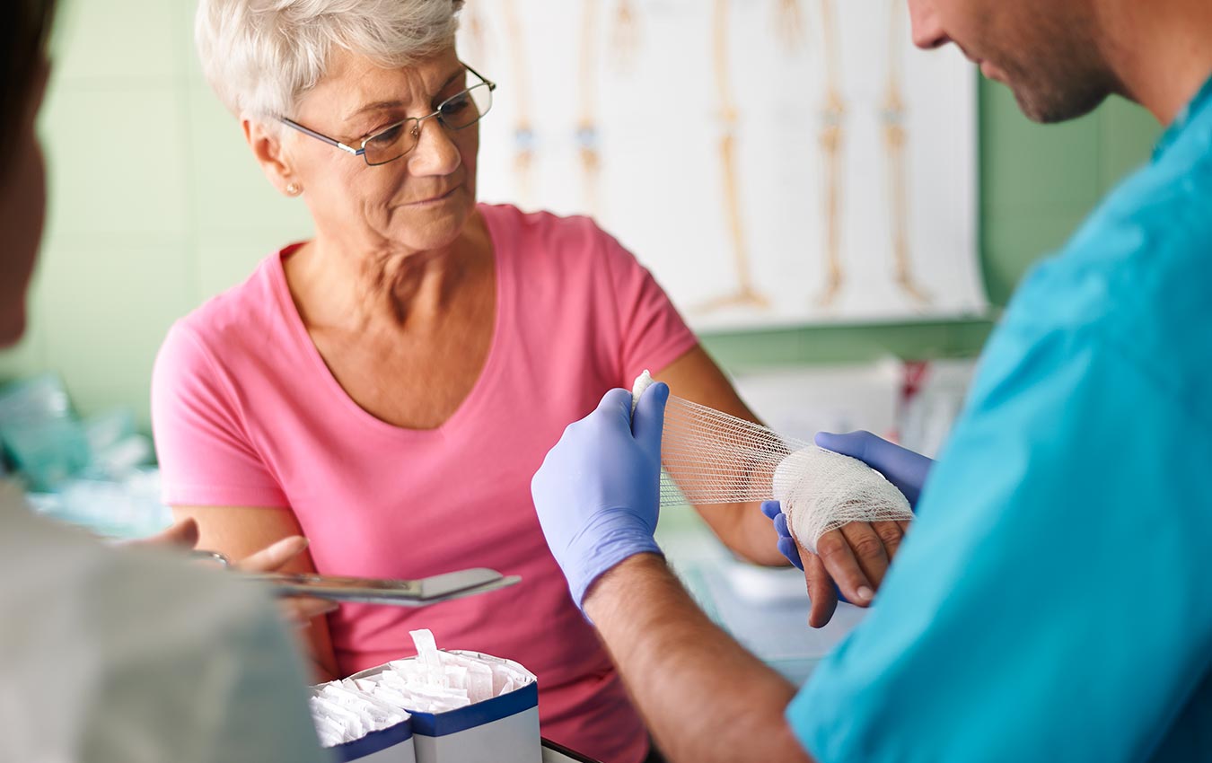 A male nurse caring for a senior woman by wrapping a bandage on her injured hand.