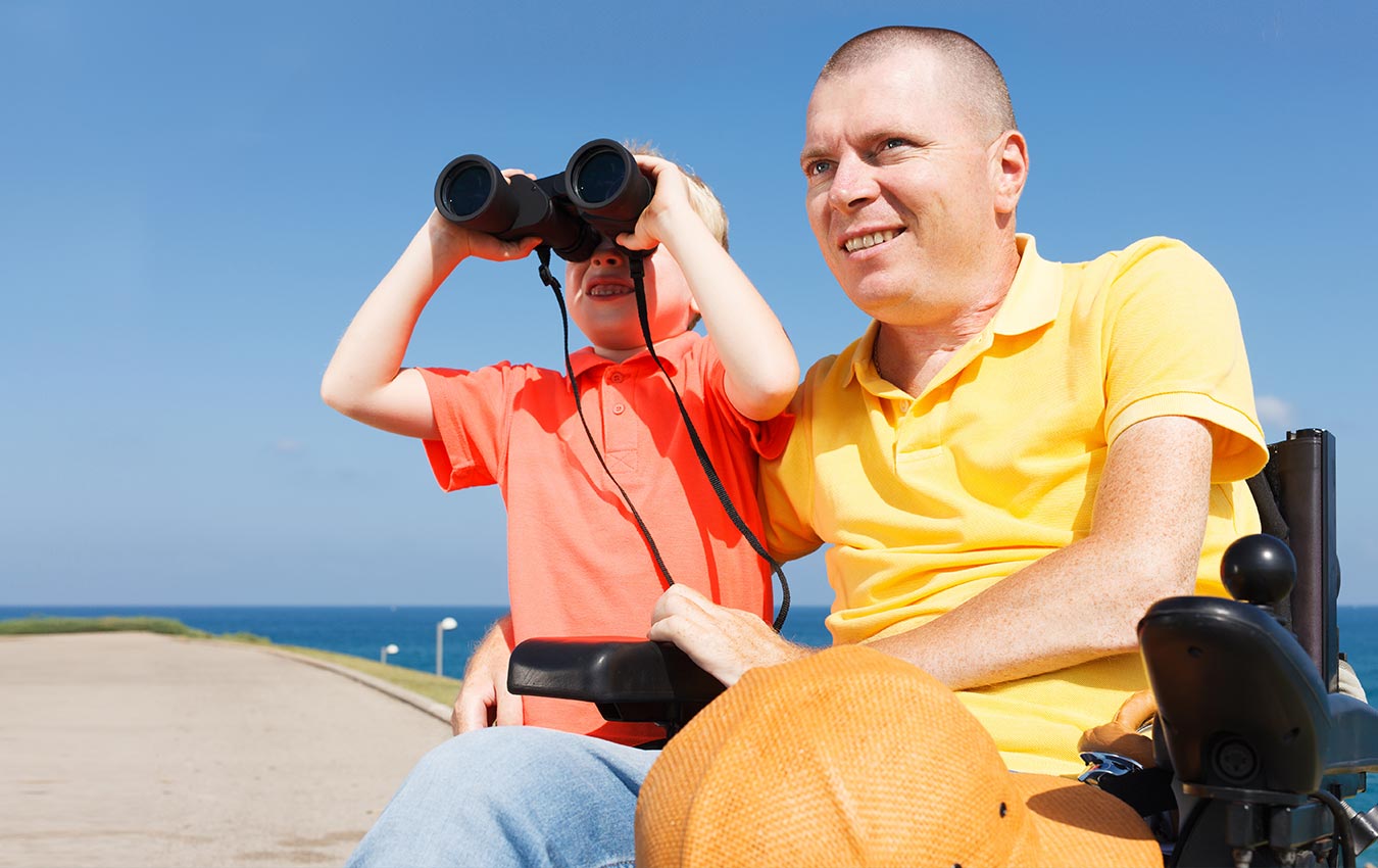 A man sitting in a walker holding his young son who is using a pair of binoculars.