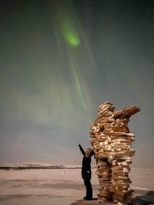 Person standing in coat in front of Inukshuk looking at northern lights