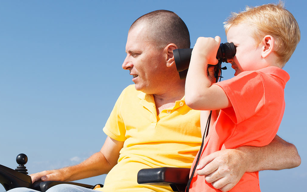 A man sat on a chair holding his son who is stood besides him. The son is using a pair of binoculars.