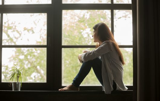 Woman sitting on windowsill embracing knees and looking outside