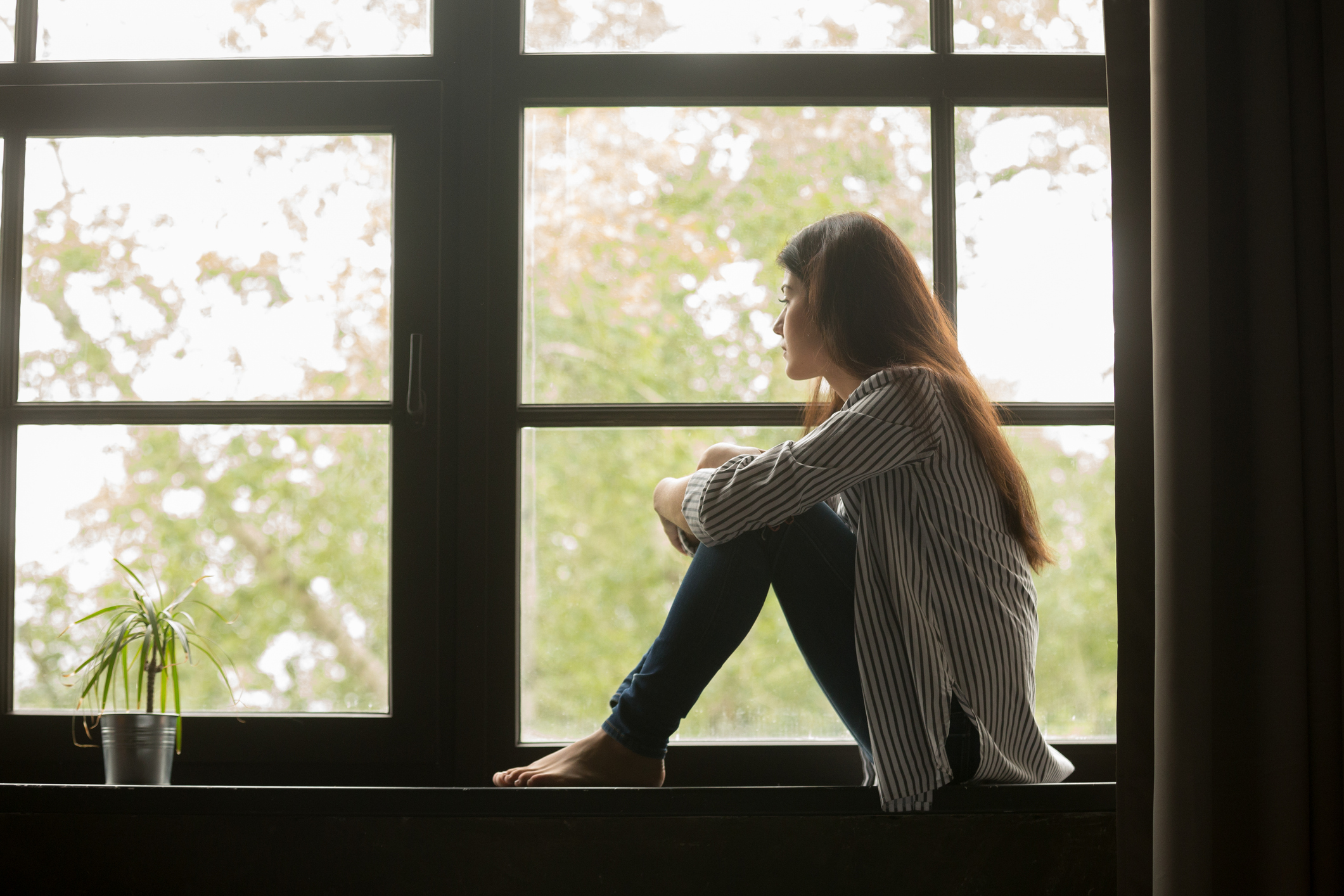 Woman sitting on windowsill embracing knees and looking outside
