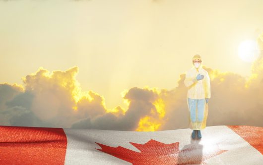 Medical worker in personal protective gear standing on Canadian flag with hand to chest