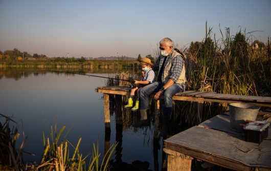 Homme senior assis sur une jetée pêchant dans le lac avec son petit-fils sous des masques covid