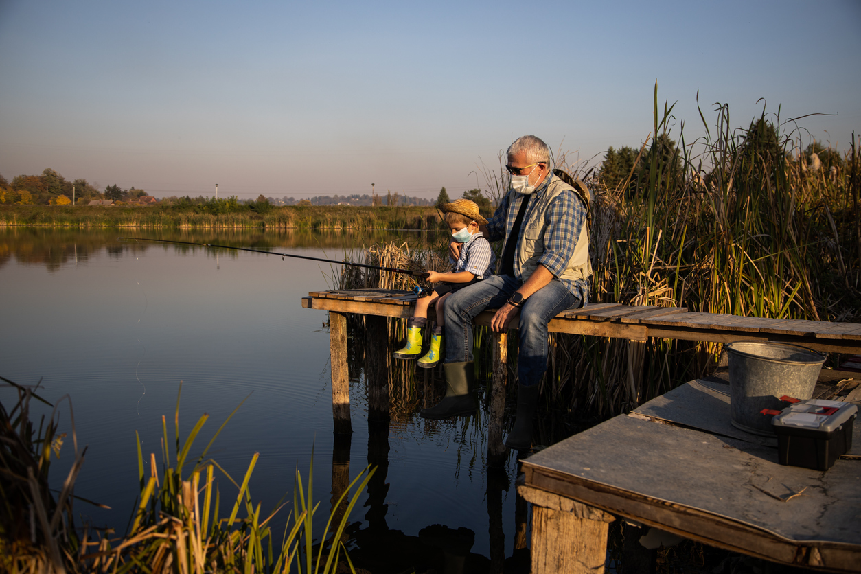 Senior man sitting on jetty fishing in lake with grandson under covid masks