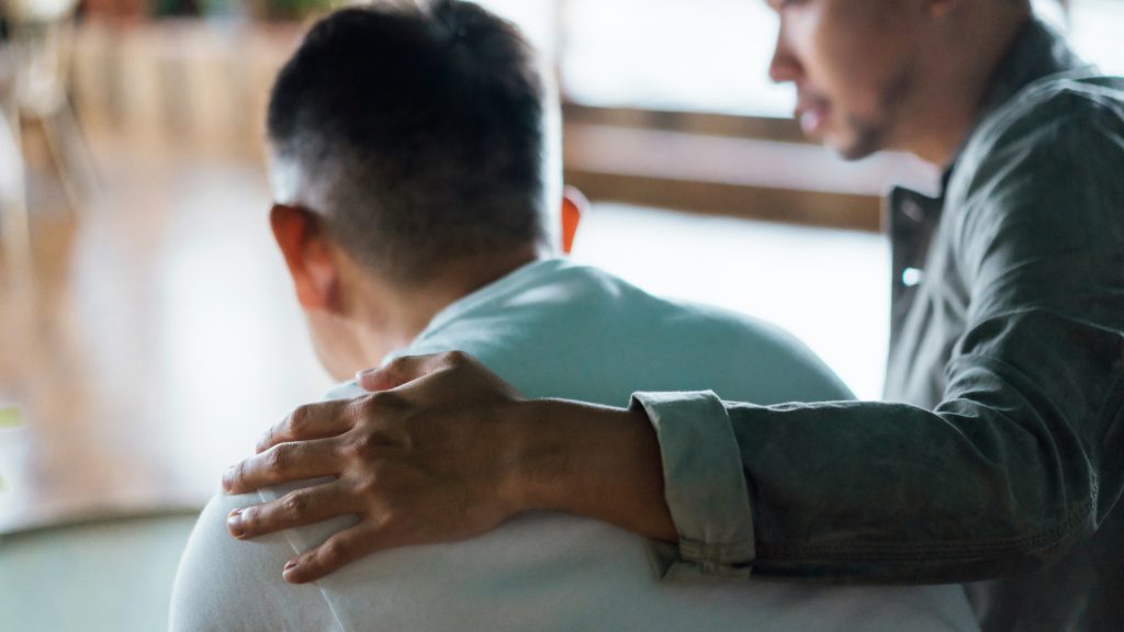 Rear view of son and elderly father sitting together at home. Son caring for his father, putting hand on his shoulder, comforting and consoling him.