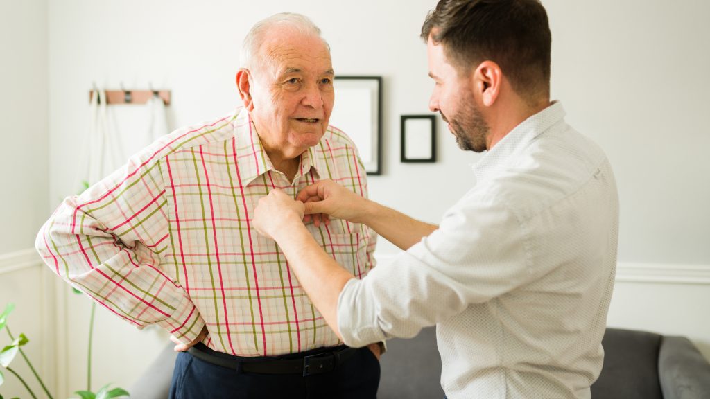 Caring young son helping his old happy elderly father to change clothes before going out of the house together