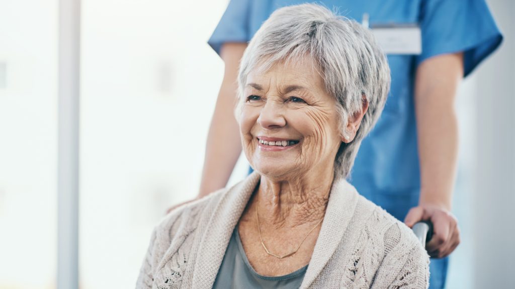 Happy and excited senior woman sitting in a wheelchair being pushed by her caregiver. Mature or old female smiling and joyful about being discharged from hospital and leaving with a nurse