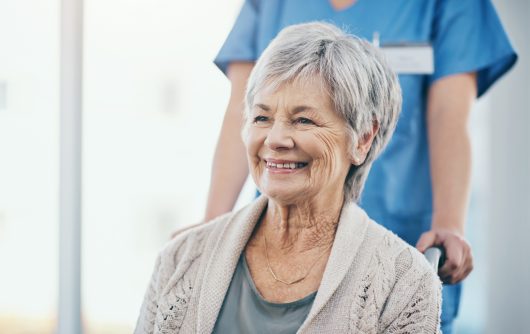 Happy and excited senior woman sitting in a wheelchair being pushed by her caregiver. Mature or old female smiling and joyful about being discharged from hospital and leaving with a nurse