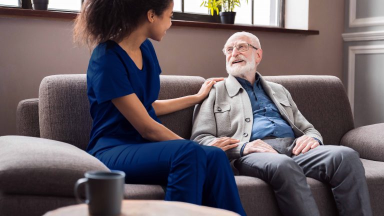 A female nurse and an elderly man sit on a couch while talking to each other and smiling.