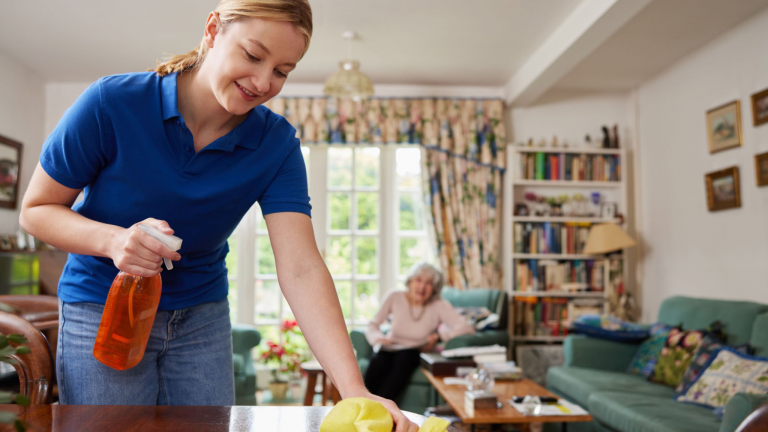 A young woman uses a spray and a towel to clean a table while an elderly woman in the background watches from a chair.