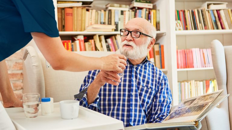 A nurse hands an elderly man reading a large book a cup of medicine.