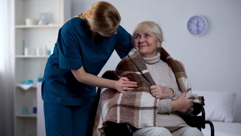 A female nurse places a blanket around the shoulders of an elderly woman in a wheelchair.