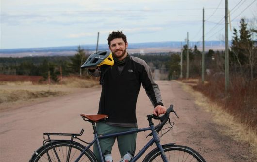 Simeon King, a type 1 diabetes advocate, stands on a rural road with his touring bicycle, holding a bike helmet and smiling at the camera. He is preparing for his coast-to-coast "Cycle for Type 1" ride across Canada to raise awareness and funds for I Challenge Diabetes.