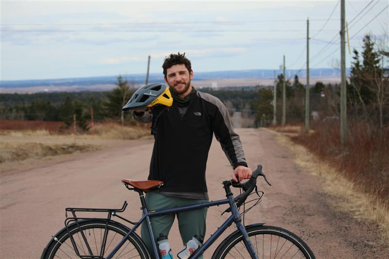 Simeon King, a type 1 diabetes advocate, stands on a rural road with his touring bicycle, holding a bike helmet and smiling at the camera. He is preparing for his coast-to-coast "Cycle for Type 1" ride across Canada to raise awareness and funds for I Challenge Diabetes.