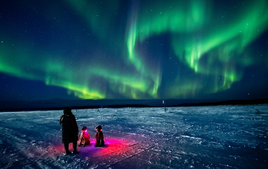 A trio on a snowy landscape under vibrant green Northern Lights. The ground glows with a soft red light, creating a serene and awe-inspiring atmosphere.