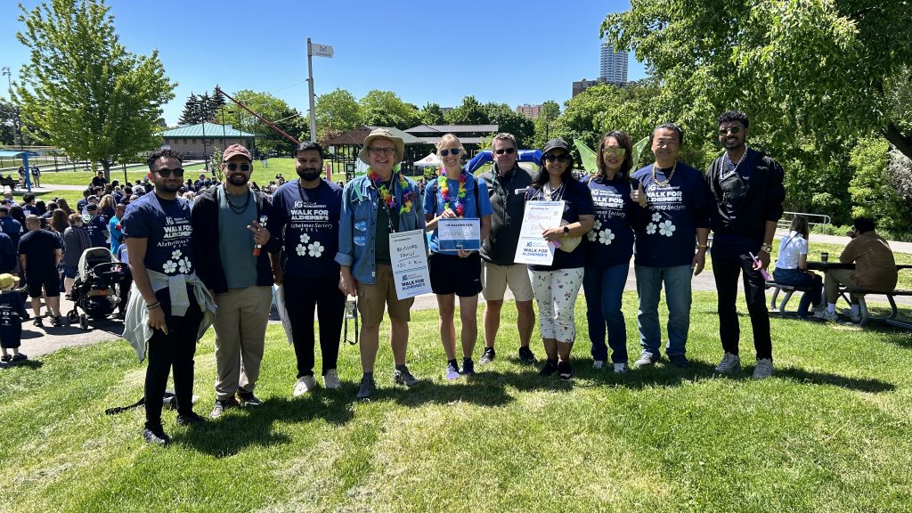 A group of ten people stands in a line on a grassy hill on a sunny day, posing for a photo at an IG Wealth Management Walk for Alzheimer's event.