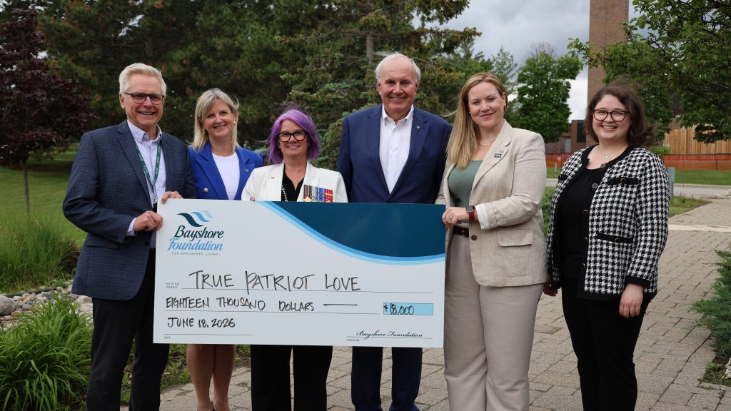 Six people standing outdoors holding a large ceremonial check from the Bayshore Foundation for $18,000, made out to True Patriot Love and dated June 18, 2026.