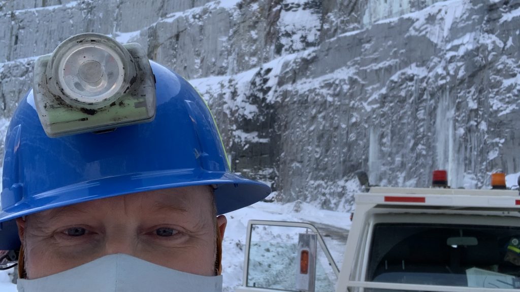 Close-up selfie of a worker wearing a blue hard hat, face mask, and high-vis jacket, standing near an ambulance at an icy mine or quarry.