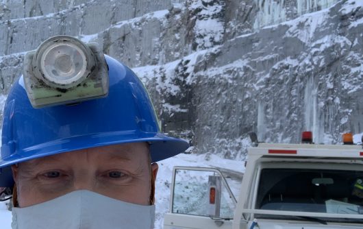 Close-up selfie of a worker wearing a blue hard hat, face mask, and high-vis jacket, standing near an ambulance at an icy mine or quarry.