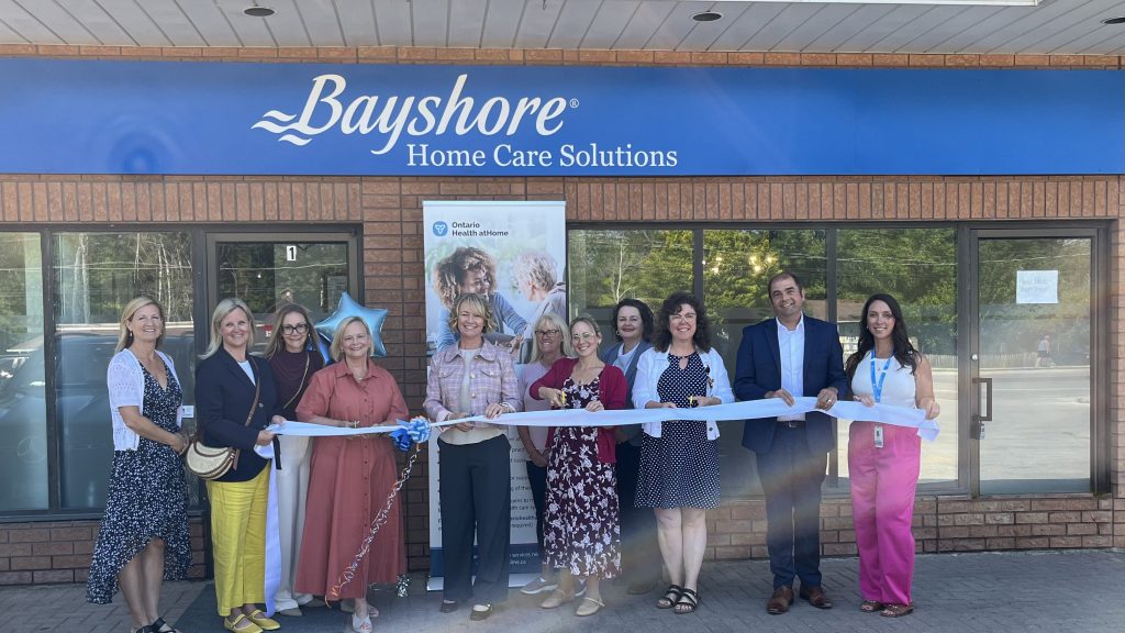 A group of ten people participates in a ribbon-cutting ceremony outside a Bayshore Home Care Solutions building next to an Ontario Health at Home banner.