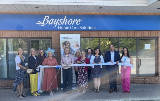 A group of ten people participates in a ribbon-cutting ceremony outside a Bayshore Home Care Solutions building next to an Ontario Health at Home banner.