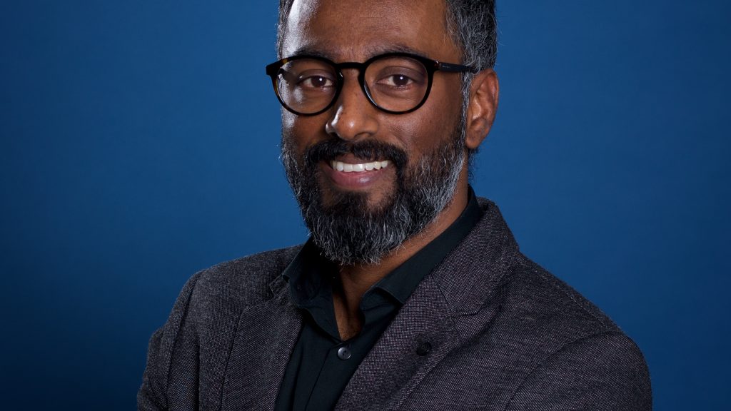Une photo de profil professionnelle de Dheeraj Paul, un homme souriant avec des lunettes, des cheveux courts foncés et une barbe fournie.