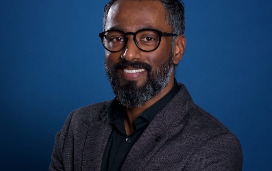 Une photo de profil professionnelle de Dheeraj Paul, un homme souriant avec des lunettes, des cheveux courts foncés et une barbe fournie.