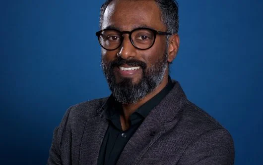A professional headshot of Dheeraj Paul, a smiling man with glasses, short dark hair, and a full beard.