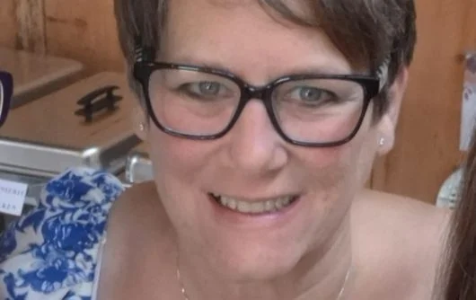 A woman with short brown hair, dark-rimmed glasses, and a thin necklace smiling at the camera. She is wearing a blue and white patterned top and standing in front of a wooden panel background.