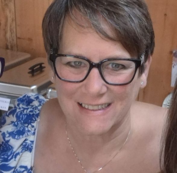 A woman with short brown hair, dark-rimmed glasses, and a thin necklace smiling at the camera. She is wearing a blue and white patterned top and standing in front of a wooden panel background.